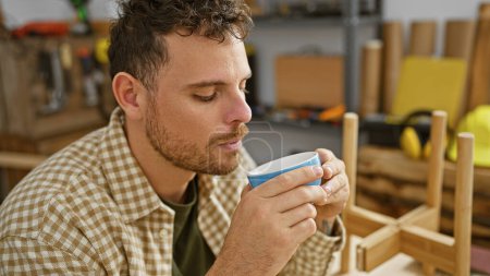 Bearded man enjoys coffee break in woodworking studio, capturing his casual style and intimate craftsmanship ambiance.