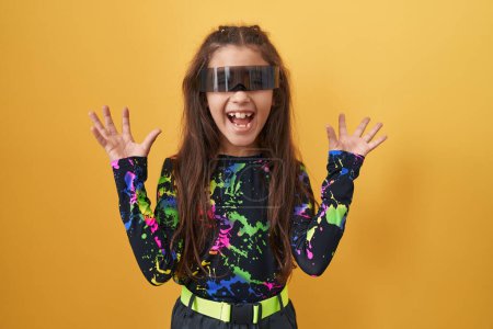 Téléchargez les photos : Adorable fille hispanique, folle de joie, célèbre excitée victoire de la réalité virtuelle, bras levés, lunettes sur. triomphe personnifié sur fond jaune isolé - en image libre de droit