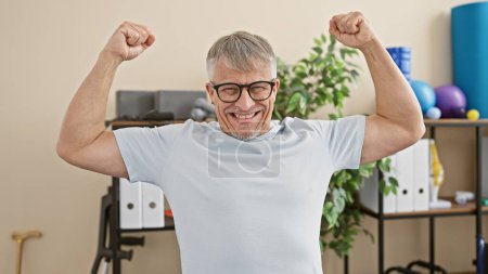 Foto de Hombre mayor sonriendo y flexionando los músculos con confianza en un centro de rehabilitación. - Imagen libre de derechos
