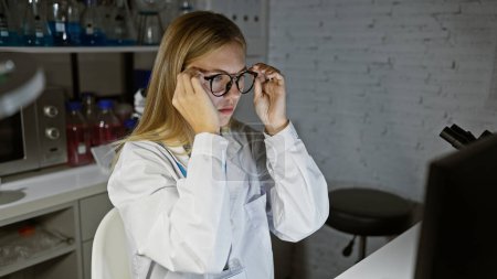 Foto de Mujer rubia ajustando gafas en entorno de laboratorio, retratando la inteligencia y la precisión. - Imagen libre de derechos