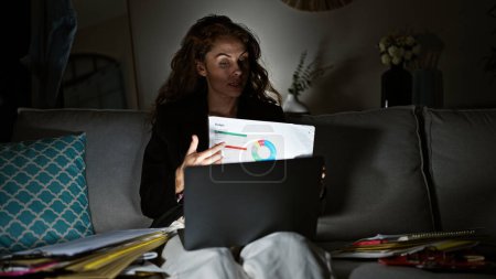 A focused caucasian woman analyzes financial charts on paper while working late on her laptop in a dimly lit living room.