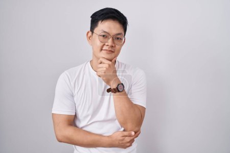 Photo for Young asian man standing over white background looking confident at the camera smiling with crossed arms and hand raised on chin. thinking positive. - Royalty Free Image