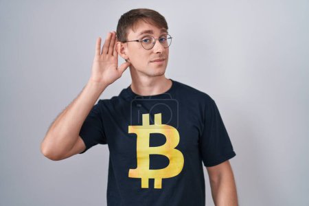 Photo for Caucasian blond man wearing bitcoin t shirt smiling with hand over ear listening an hearing to rumor or gossip. deafness concept. - Royalty Free Image