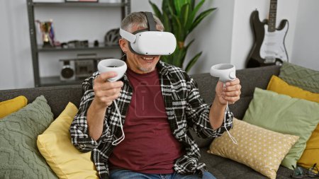 Foto de Un hombre alegre de mediana edad experimenta la realidad virtual con auriculares y controladores vr en una acogedora sala de estar, expresando emoción. - Imagen libre de derechos