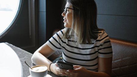 Foto de Mujer hispana joven disfrutando del café y el teléfono inteligente en un interior moderno café - Imagen libre de derechos