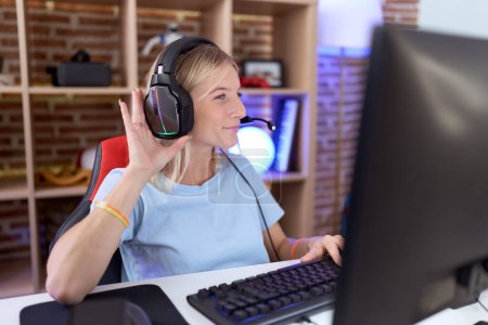 Photo for Young caucasian woman playing video games wearing headphones smiling with hand over ear listening an hearing to rumor or gossip. deafness concept. - Royalty Free Image