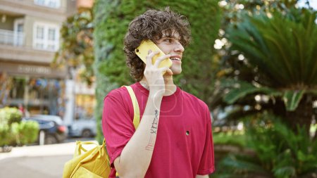 A curly-haired young man, holding a yellow phone to his ear, smiles in an urban park setting with green foliage background.