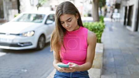 Foto de Atractiva mujer hispana joven, inmersa en su propio mundo digital, se sienta relajada pero centrada en un banco de la calle de la ciudad, usando su teléfono inteligente, comprometida en una conversación seria de texto. - Imagen libre de derechos