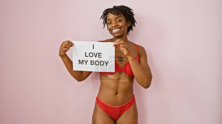 Téléchargez les photos : Une jeune femme noire confiante avec des dreadlocks pose en lingerie rouge sur un fond rose, tenant un signe "j'aime mon corps". - en image libre de droit
