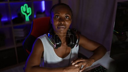 Téléchargez les photos : Femme afro-américaine focalisée avec casque de jeu à la maison la nuit, mettant en valeur la technologie et le divertissement. - en image libre de droit