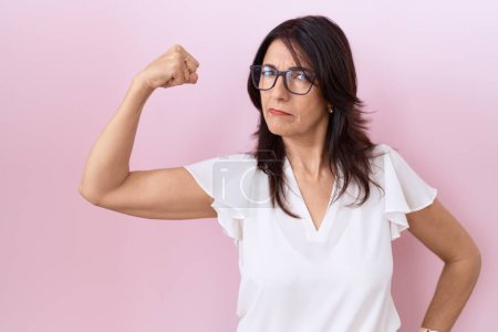 Foto de Mujer hispana de mediana edad vistiendo camiseta blanca casual y gafas persona fuerte mostrando músculo del brazo, confiado y orgulloso de poder - Imagen libre de derechos