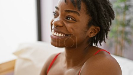 Téléchargez les photos : Une jeune femme noire souriante avec dreadlocks, vêtue d'un haut rouge, assise dans une chambre lumineuse avec un fond flou. - en image libre de droit