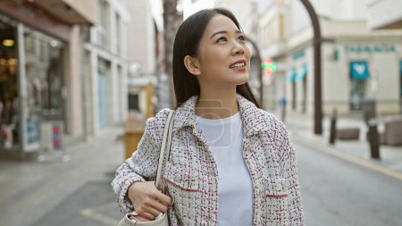 Téléchargez les photos : Une jeune femme asiatique vêtue de façon décontractée se promène avec un sac dans une rue ensoleillée de la ville qui dégage un sentiment d'exploration et de confort. - en image libre de droit