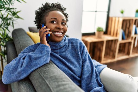 Foto de Mujer afroamericana hablando en el teléfono inteligente sentado en el sofá en casa - Imagen libre de derechos