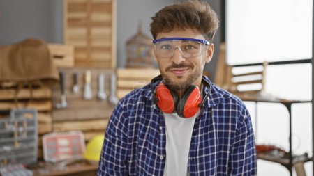 Téléchargez les photos : Un jeune homme arabe souriant avec des lunettes debout avec confiance dans son atelier de menuiserie, embrassant la joie du travail du bois. - en image libre de droit