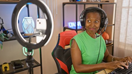 Téléchargez les photos : Femme afro-américaine avec écouteurs dans une salle de jeux à domicile à l'aide d'un ordinateur et souriant - en image libre de droit