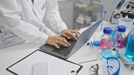 Photo for Hispanic woman scientist's hands deftly typing, immersed in groundbreaking medical research, working indoors at a bustling lab using laptop computer. - Royalty Free Image