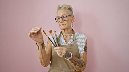 Photo for Serious-faced, grey-haired senior woman artist intensely holding paintbrushes over pink, isolated background - a painting class in full swing - Royalty Free Image