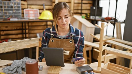 Photo for Attractive young blonde female carpenter, engrossed in serious business of financing her carpentry workshop, using credit card & touchpad amidst timber, indoor - Royalty Free Image