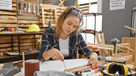 Attractive young hispanic woman carpenter, dressed for safety with glasses, skillfully jots down notes in her carpentry workshop