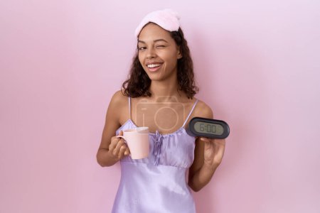 Photo for Young hispanic woman wearing nightgown holding alarm clock winking looking at the camera with sexy expression, cheerful and happy face. - Royalty Free Image