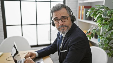 Smiling young hispanic man, a grey-haired business worker, buzzes with success as he handles business on his laptop, headphones on, in a lively office.