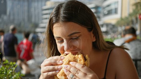 Photo for A young hispanic woman enjoying a sandwich outdoors with skyscrapers, including the burj khalifa, in dubai. - Royalty Free Image