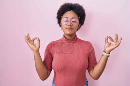 Téléchargez les photos : Belle femme africaine aux cheveux bouclés debout sur fond rose détendue et souriante avec les yeux fermés faisant geste de méditation avec les doigts. concept de yoga. - en image libre de droit