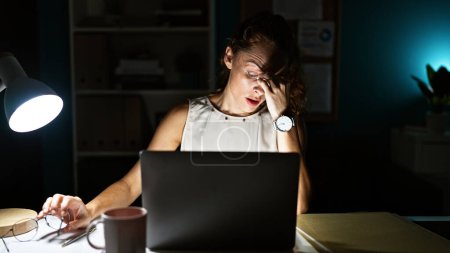 Foto de Una joven parece estresada trabajando hasta tarde en su oficina en un portátil, iluminando su cara con una lámpara de escritorio. - Imagen libre de derechos