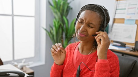Foto de Mujer afroamericana en un entorno de oficina con auriculares con una expresión problemática - Imagen libre de derechos