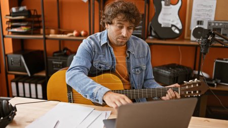 Photo for Handsome hispanic man playing guitar in a music studio while looking at a laptop. - Royalty Free Image