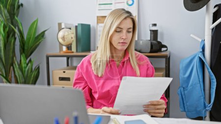 Photo for A focused caucasian woman in a vibrant pink shirt reads documents in a cluttered office setting. - Royalty Free Image
