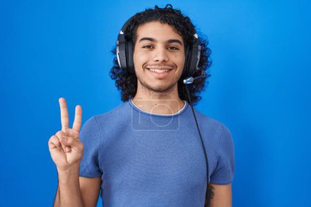 Téléchargez les photos : Homme hispanique aux cheveux bouclés écoutant de la musique à l'aide d'écouteurs montrant et pointant vers le haut avec les doigts numéro deux tout en souriant confiant et heureux. - en image libre de droit