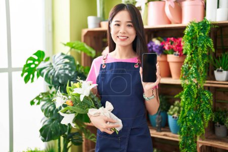 Foto de Joven mujer china que trabaja en la floristería mostrando la pantalla del teléfono inteligente sonriendo y riendo en voz alta porque divertida broma loca. - Imagen libre de derechos