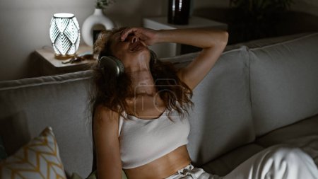 Foto de Una mujer joven disfrutando de la música con auriculares mientras se relaja en un sofá en un acogedor interior de la casa. - Imagen libre de derechos