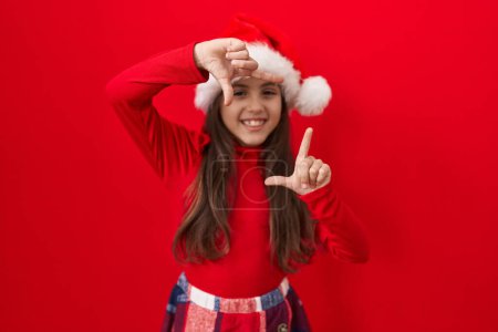 Photo for Adorable hispanic girl in a christmas hat, making a frame with hands and fingers, smiles happily over isolated red background, envisioning a photo composition. creativity and photography concept. - Royalty Free Image