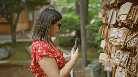Photo for Beautiful hispanic woman in glasses capturing ema prayer boards at japanese gotokuji temple, preserving tradition through a smartphone lens - Royalty Free Image