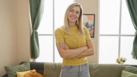 Selbstbewusste blonde Frau steht mit verschränkten Armen in einem modernen Wohnzimmer