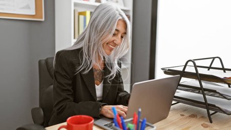 Mujer de mediana edad, confiada, sonriente, de cabello gris, disfrutando de su trabajo en un elegante escritorio ejecutivo, en su computadora portátil, irradiando éxito como trabajadora profesional en la oficina.