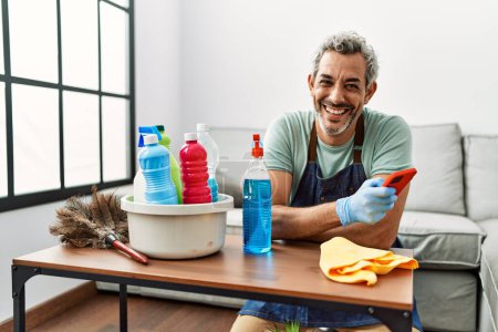 Foto de Middle age grey-haired man using smartphone cleaning table at home - Imagen libre de derechos