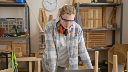 Téléchargez les photos : Focalisée femme blonde portant des lunettes de sécurité travaille à la menuiserie banc dans l'atelier intérieur. - en image libre de droit