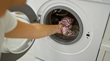 A young man doing laundry in a domestic setting, highlighting his arm and clothing inside the washing machine.