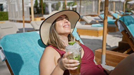 Téléchargez les photos : Une femme souriante dans un chapeau de soleil profite d'un mojito tout en se prélassant sur une chaise de plage dans un complexe de luxe. - en image libre de droit