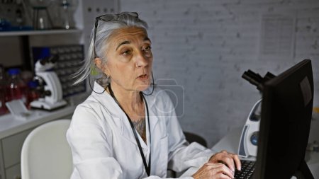 Foto de Científica de pelo gris de mediana edad, cara enfocada iluminada por el resplandor de las computadoras, trabajando diligentemente en la investigación revolucionaria en un laboratorio silencioso y débilmente iluminado - Imagen libre de derechos