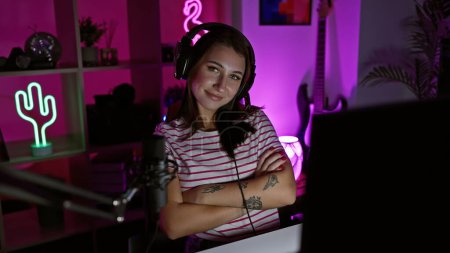 A young brunette woman with tattoos smiling, wearing headphones, in a neon-lit gaming room at night.