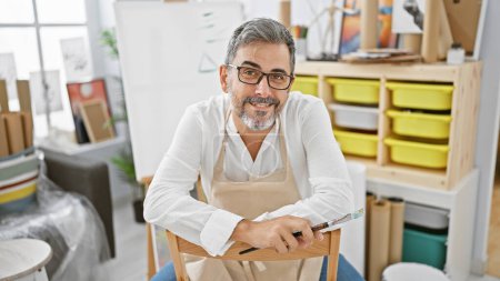 Photo for Grey-haired young hispanic man joyfully painting, a bearded artist with glasses, smiling confidently, sitting in an art studio holding a paintbrush - Royalty Free Image