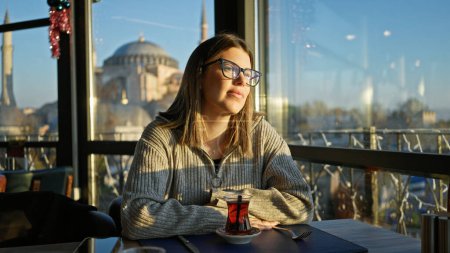 Photo for A contemplative young woman sips tea in a turkish restaurant with the hagia sophia in the background. - Royalty Free Image
