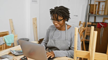 Téléchargez les photos : Une jeune femme afro-américaine avec dreadlocks travaillant sur un ordinateur portable dans un atelier de menuiserie. - en image libre de droit