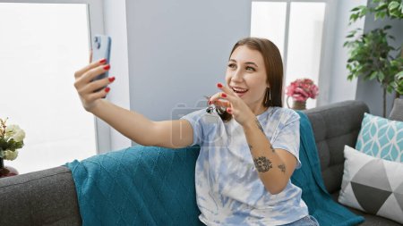 A young woman takes a selfie with keys in her cozy living room, exuding excitement and accomplishment.