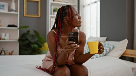Photo for Beautiful african american woman comfortably relaxed in bed, awake & typing on her smartphone, seriously concentrated, drinking coffee in her home's bedroom interior, enjoying restful morning - Royalty Free Image